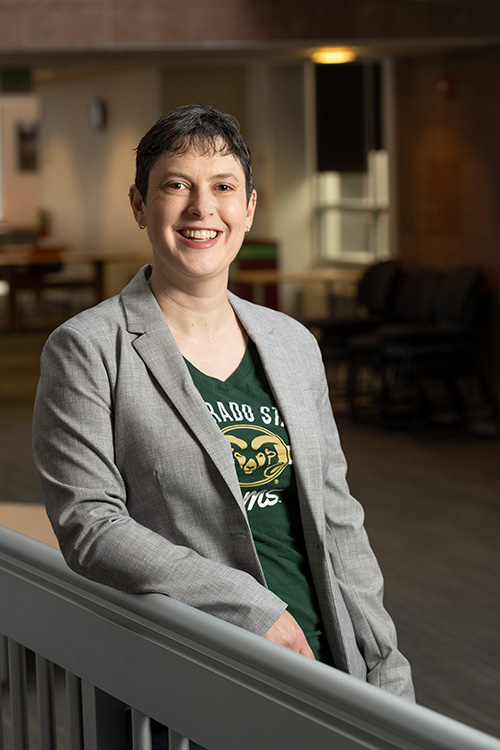 Dean of CSU Libraries Karen Estlund stands smiling at the camera. She is wearing a green and gold CSU Rams t-shirt and gray blazer. She has pale skin and short dark hair. There are library tables and chairs in the background. 