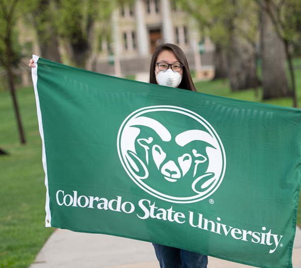 A CSU student at her graduation in 2020, wearing a protective mask and holding a flag with CSU's logo.