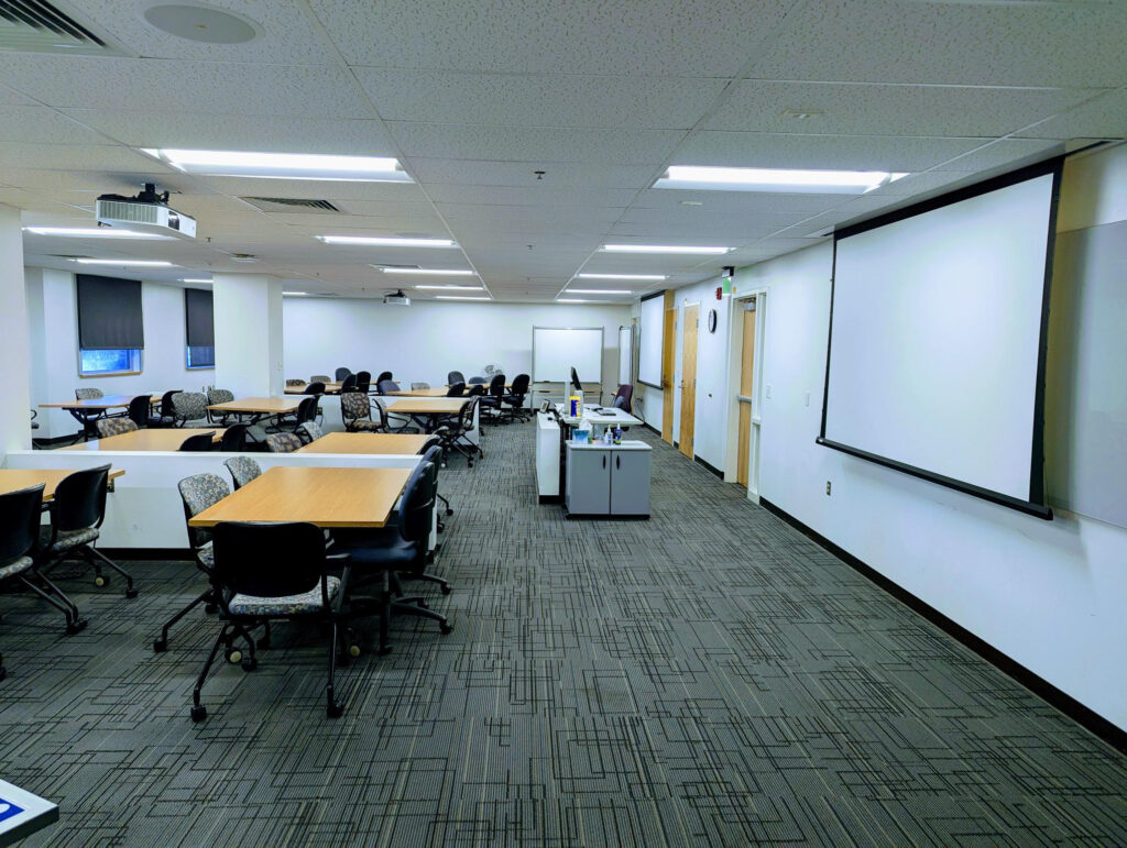 Inside of room 173, includes tables with chairs, a podium with PC, and projector with screens on either side of the room