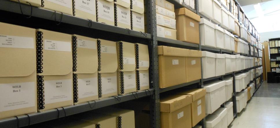 A long aisle in a storage room lined with shelves of neatly labeled archival boxes and file boxes. 