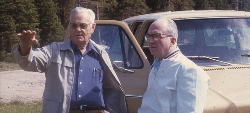 Two men clad in casual outdoor jackets discuss something off in the distance. They are standing alongside a 1970s-era vehicle on a dirt road lined with trees. 