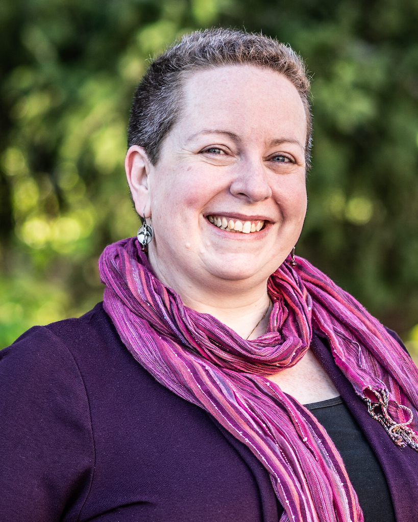 A smiling white person with short hair, wearing a scarf with greenery out of focus behind them.