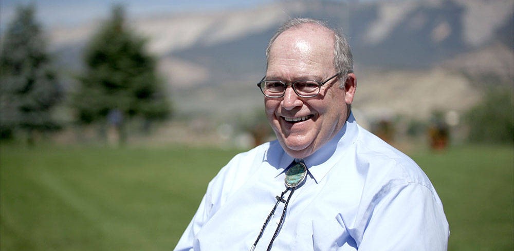 A middle-aged white man in a blue dress shirt, bolo tie, and glasses, smiling in a sunny green field with trees and mountains in the background. 