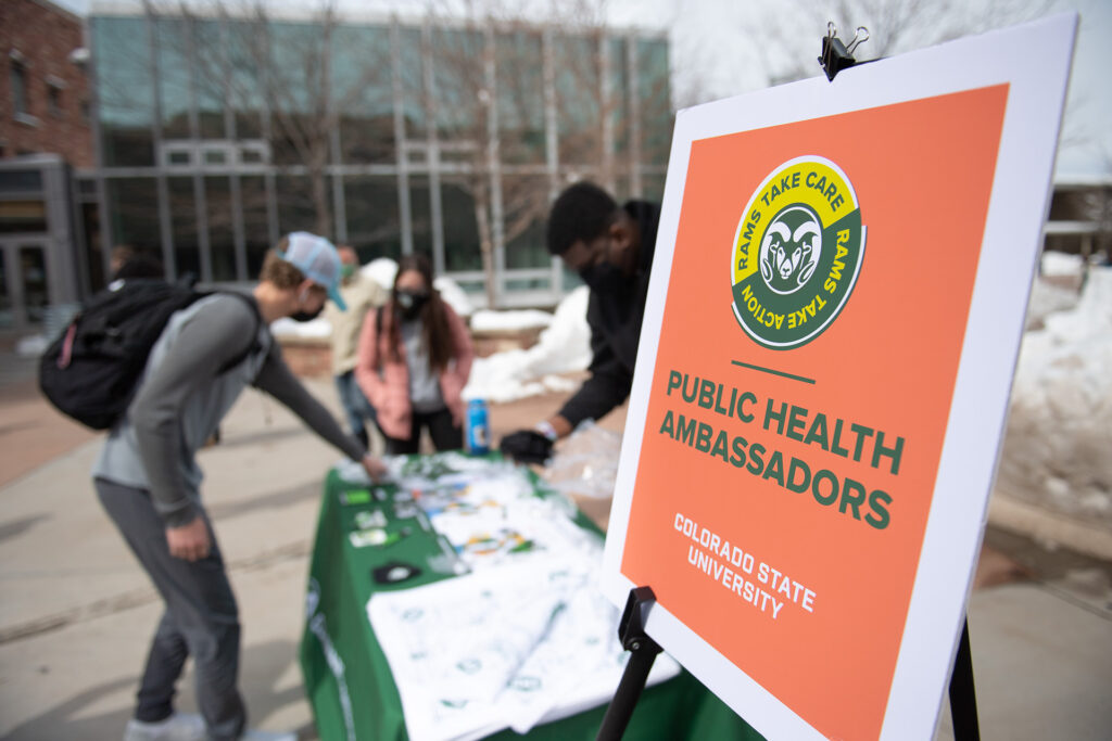 Students peruse materials at a campus outdoor tabling event next to a placard that says Public Health Ambassadors. 