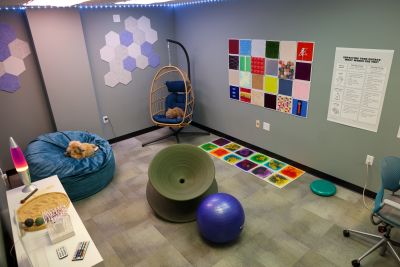 The room has calm grey walls, multiple lighting options, various textures and seating options, a sand garden, and a small collection of stims.